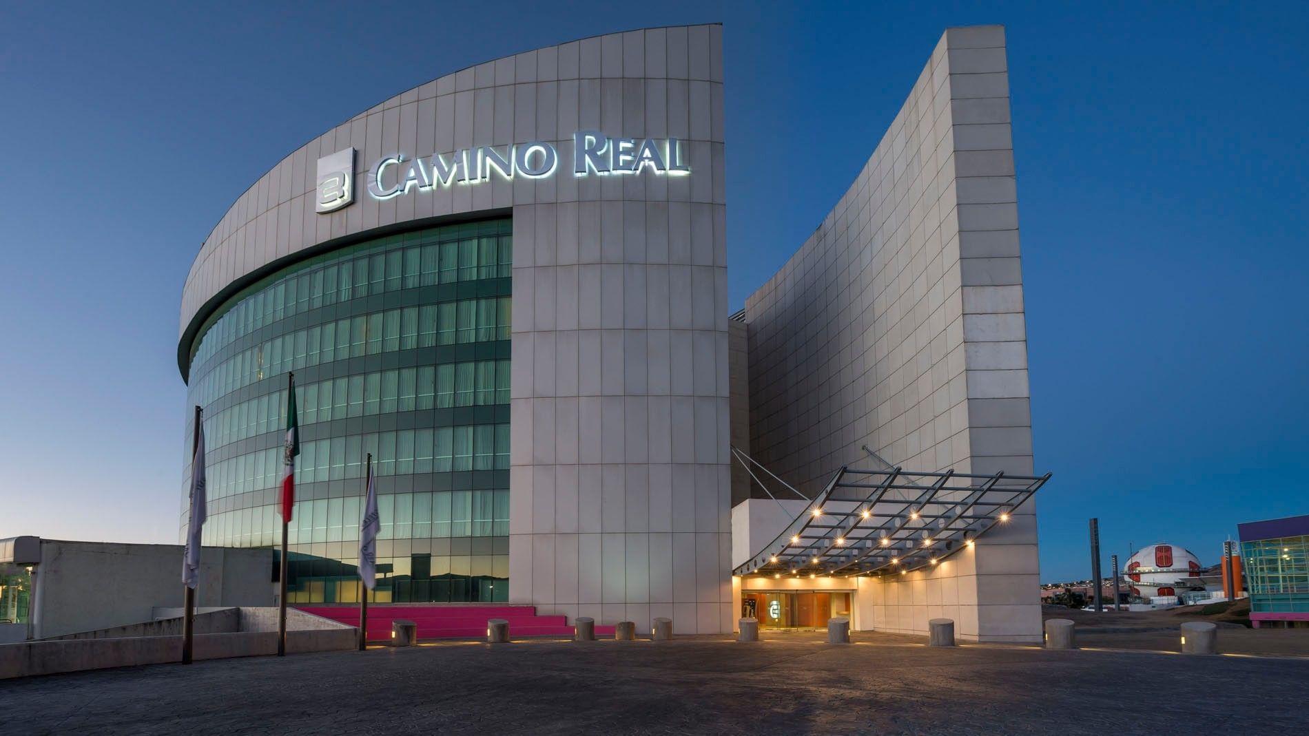 Modern curved glass exterior of Camino Real Pachuca at dusk, featuring a lit sign and bright entrance