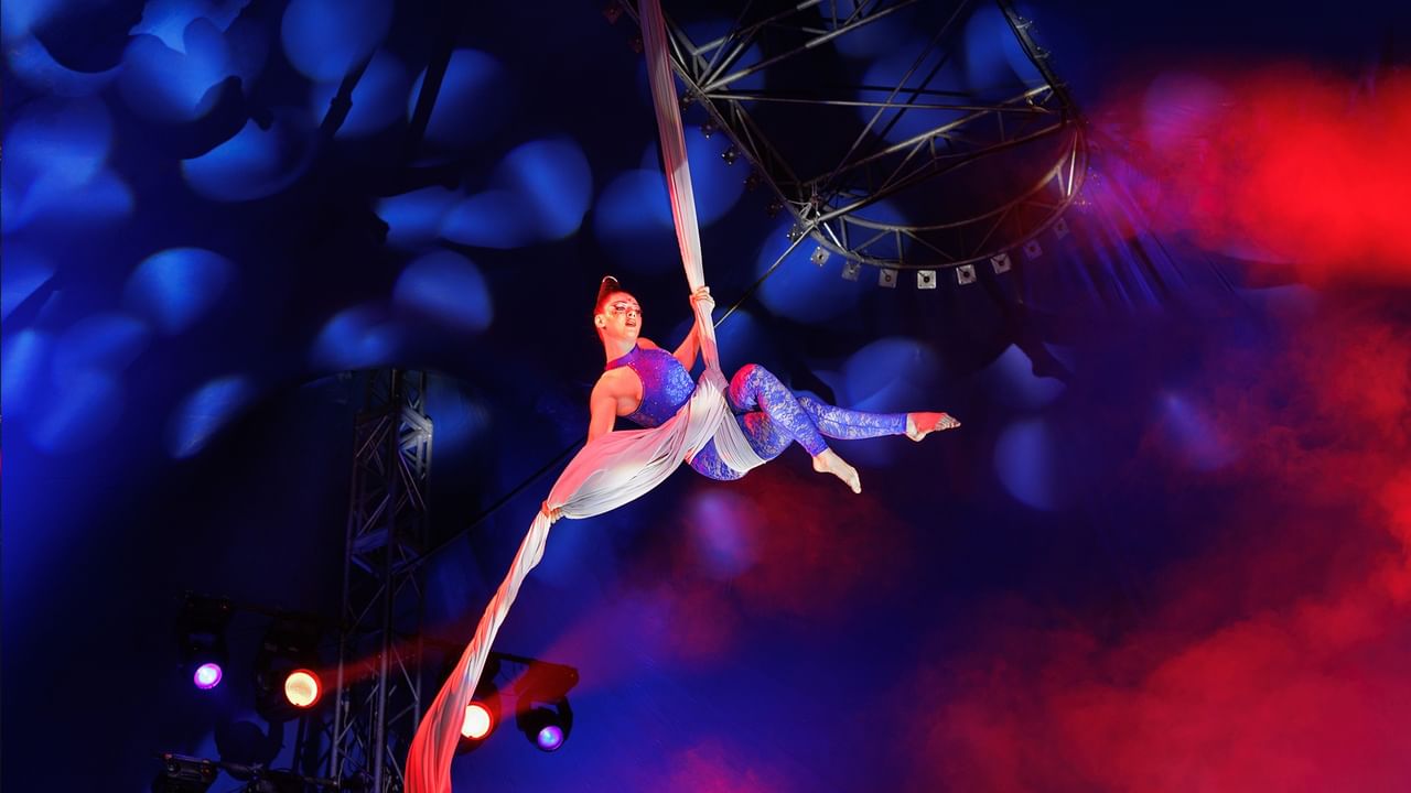 A performer in a blue outfit is suspended on a silk rope, performing acrobatics under colorful lighting.