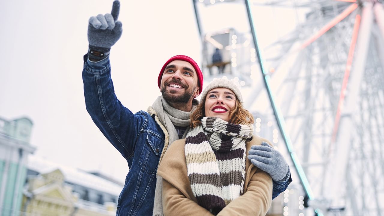 Couple in winter gear, smiling and posing for a photo in front of a Ferris wheel.