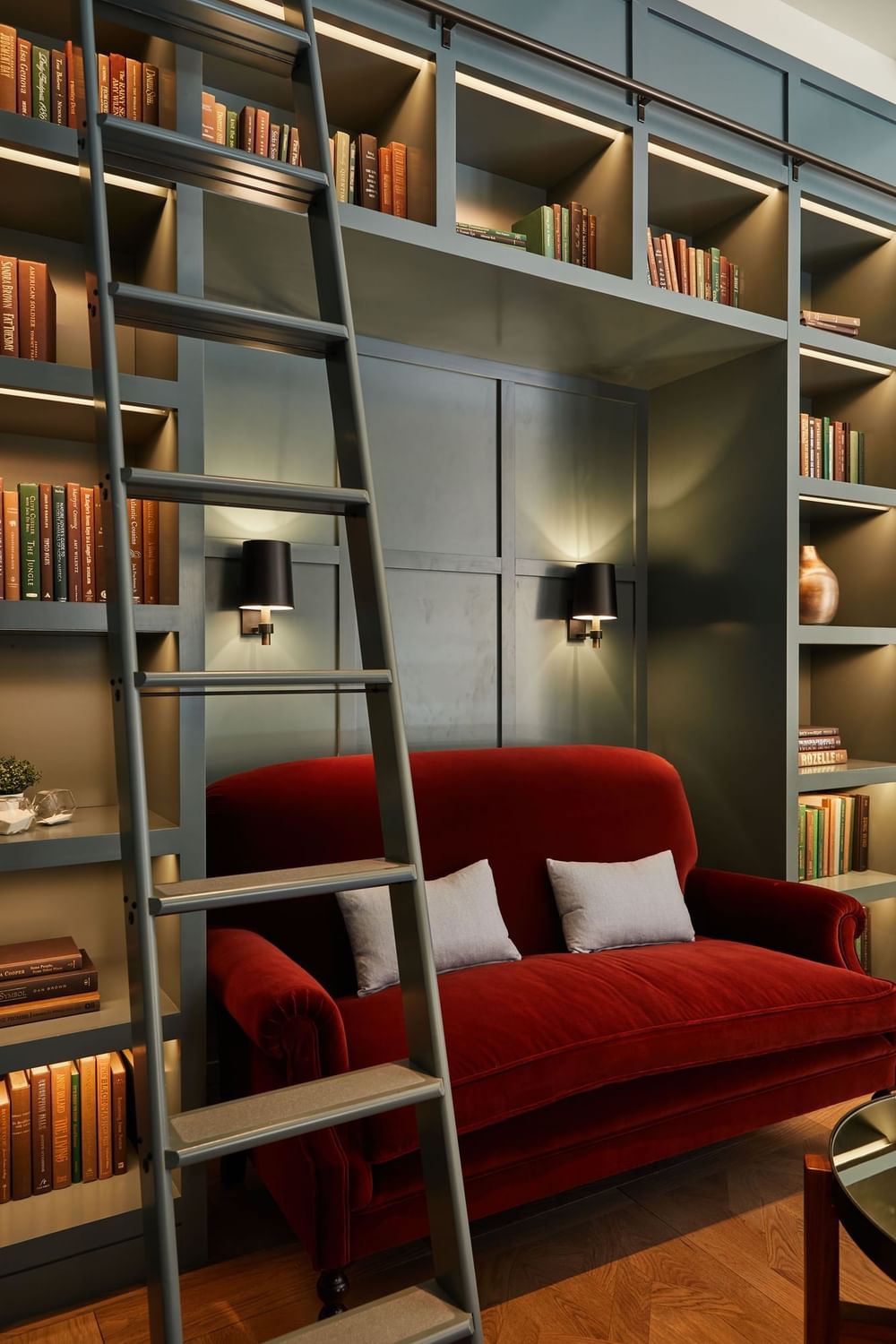 Cozy library nook featuring a red velvet sofa, bookshelves with a ladder, and soft lighting at El Prado Hotel