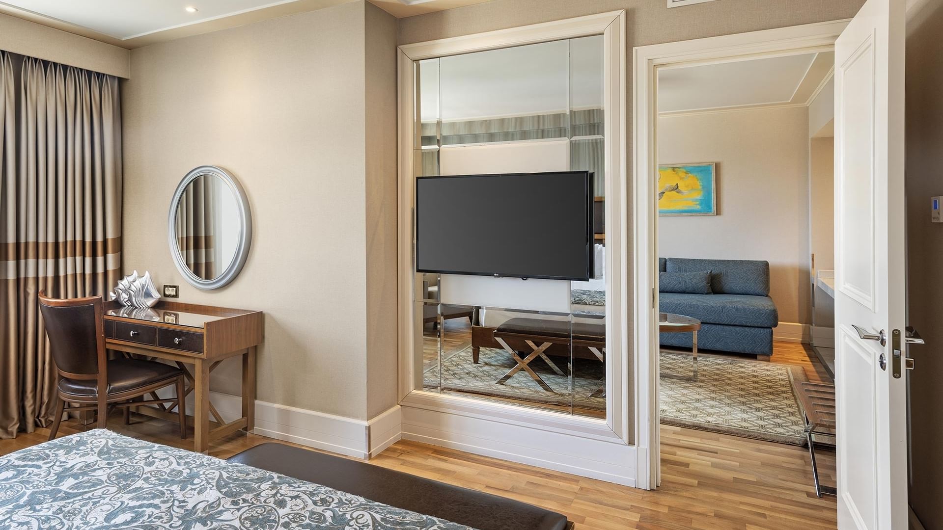 Elegant suite with bed, desk, mirror, TV, and couch visible through a mirrored glass wall at Titanic City Taksim.