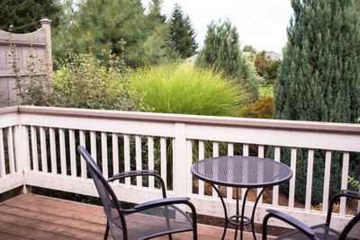 Two chairs and a table on a wooden deck with a white railing and a lush green garden view.