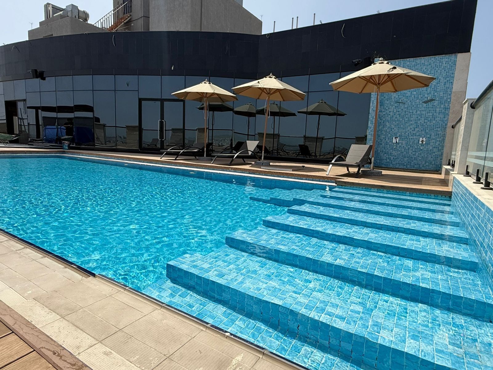 Outdoor pool with blue tiles by lounge chairs under beige umbrellas at Warwick Jeddah Hotel & Spa