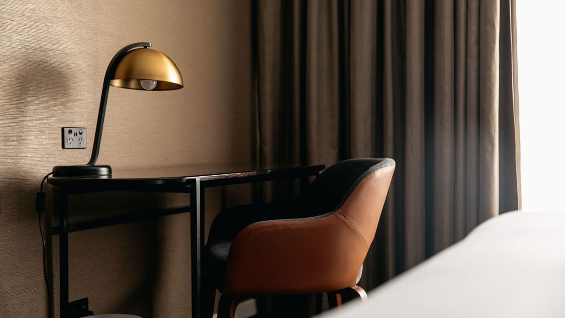 Desk with a lamp and brown chair beside a bed and curtain in a room.