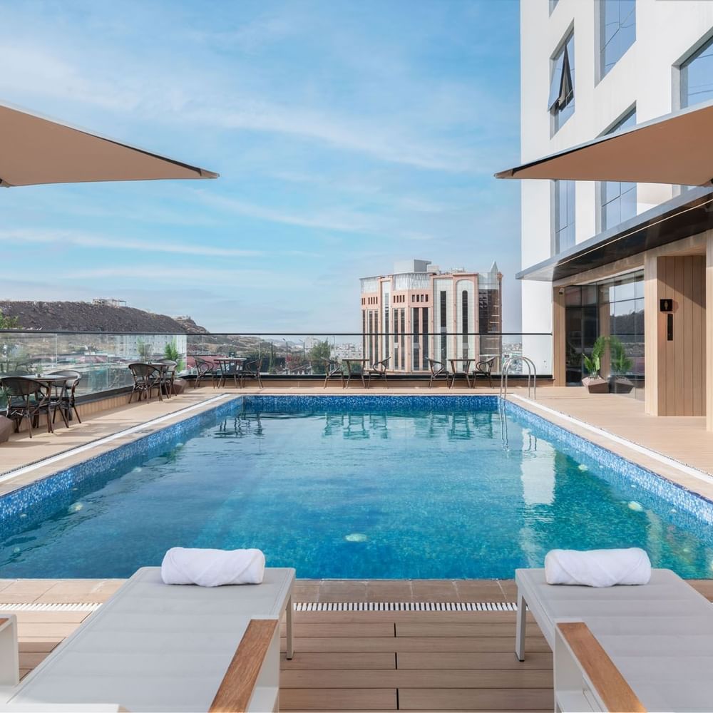 Chaises longues et tables à manger disposées au bord de la piscine sur le toit, avec vue sur la ville, à l'hôtel Warwick Al Taif