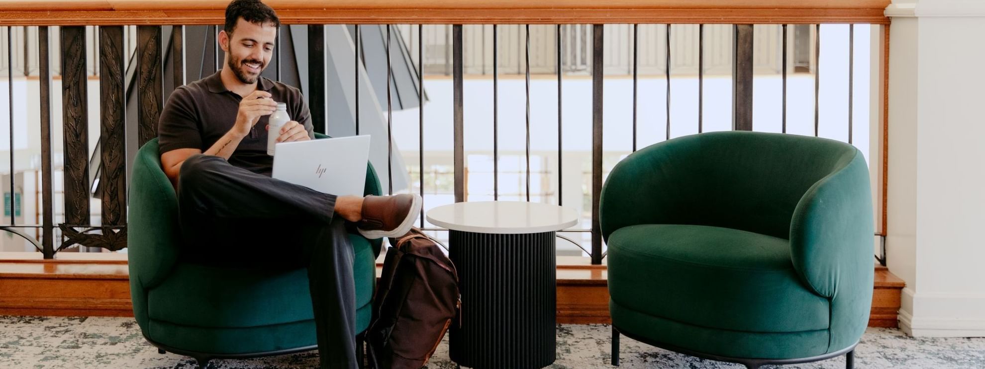 Man using laptop and drinking a beverage while sitting on green armchair next to empty armchair.