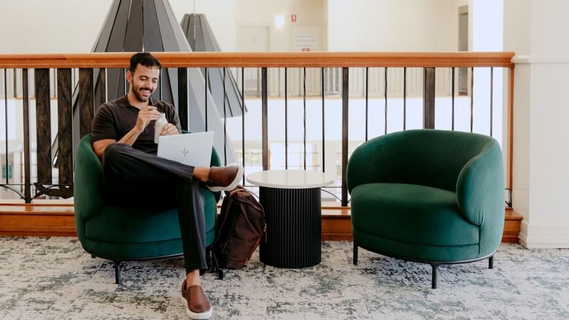 Man using laptop and drinking a beverage while sitting on green armchair next to empty armchair.