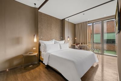 Habitación con cama grande, cojines blancos, mesa, silla, y amplias ventanas con vista a edificio de ladrillo.