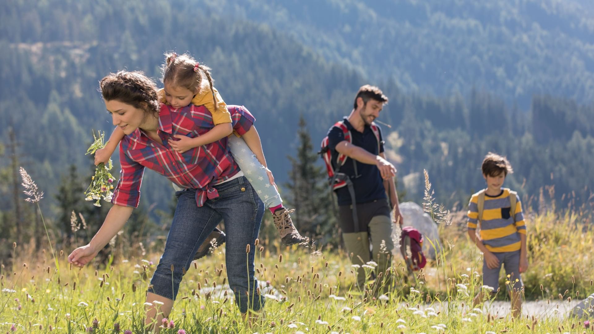 Famiglia che passeggia in montagna con bambino sulle spalle e fiori in mano at Falkensteiner