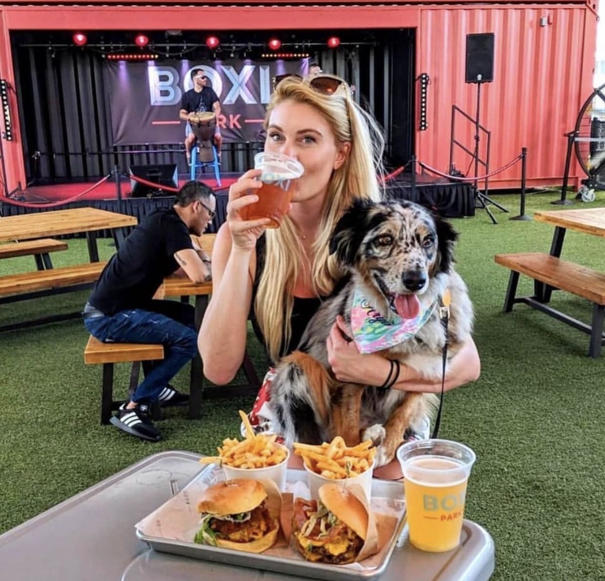 A woman sips a beer with a dog in her lap at a table in front of a stage with a backdrop that reads Boxi Park.