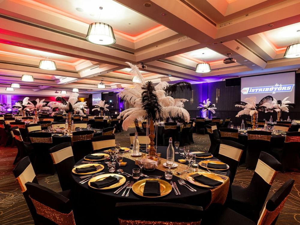 Elegant banquet hall with round tables, black chairs, gold and black decor, and a large screen displaying 