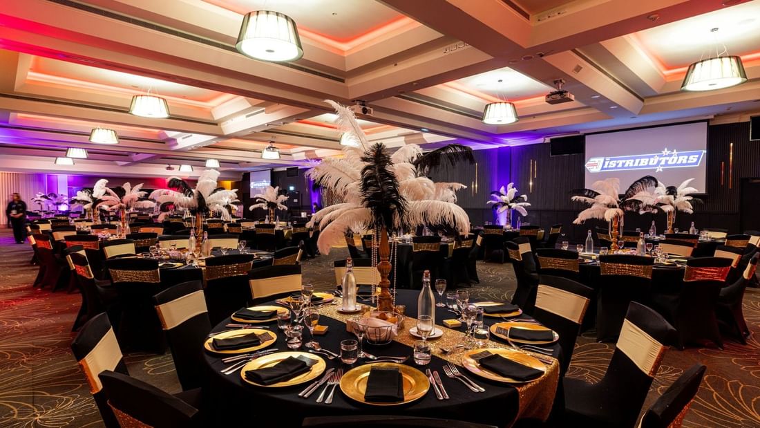 Elegantly set banquet hall with black chairs, gold and black table settings, and decorative ostrich feathers.