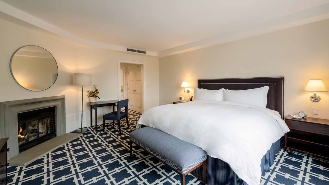 Work desk and ottoman by the cozy bed in Courtyard Deluxe Fireplace King with carpeted floors at El Prado Hotel