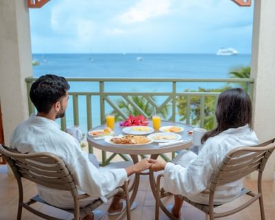 Couple in white robes enjoying a breakfast spread on a balcony with a serene ocean view at Bay Gardens Hotel
