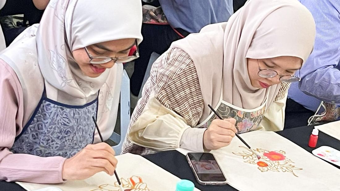 Two women wearing hijabs paint fabric at a Jadi Batek workshop.