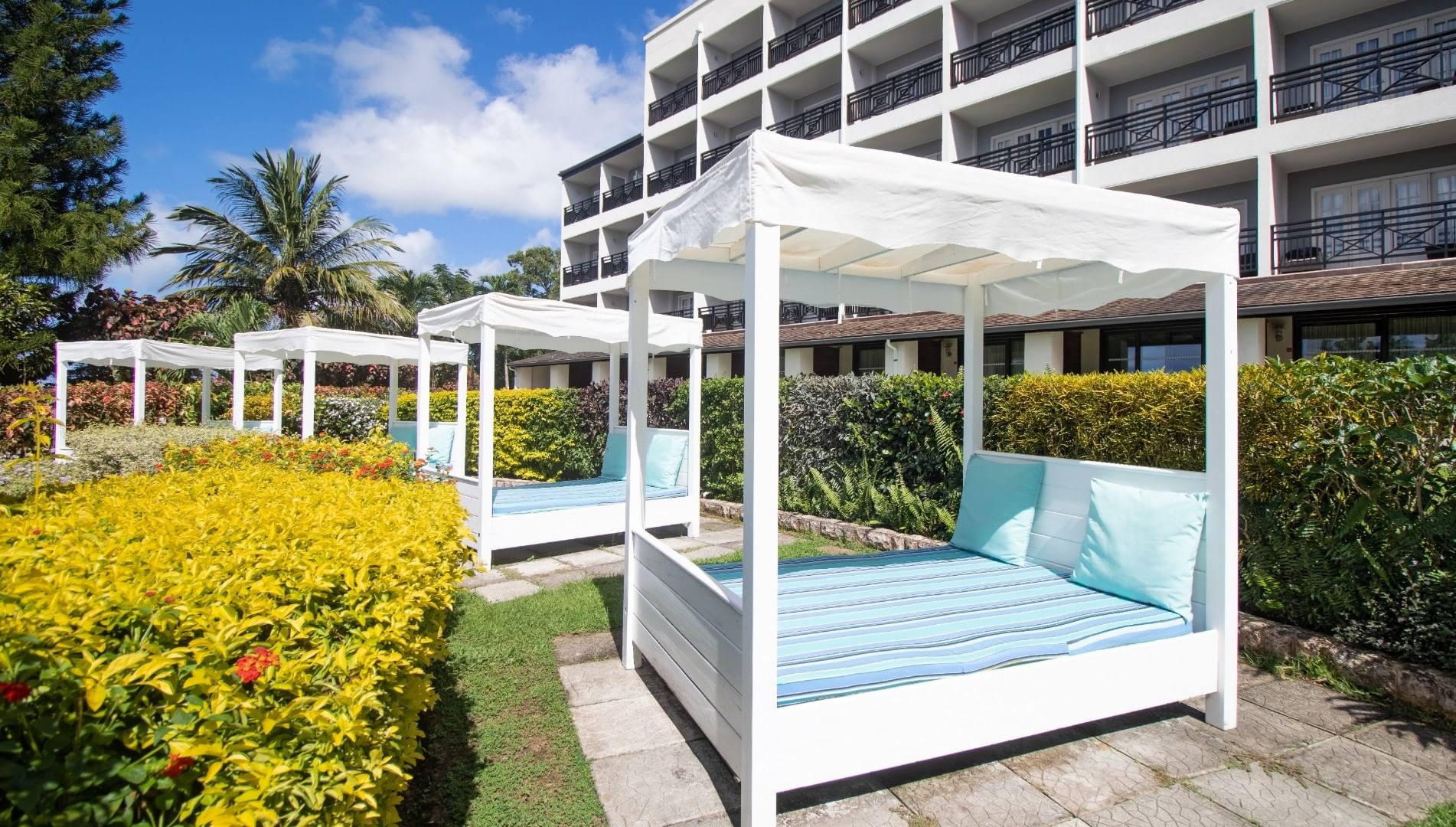 White canopy daybeds with striped blue cushions lined up on a lawn outside the Bel Jou Hotel building facade on a sunny day