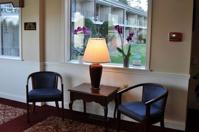 Fox Ridge Hotel waiting room with two chairs, a table with a lamp, and a window with flowers are in a room.