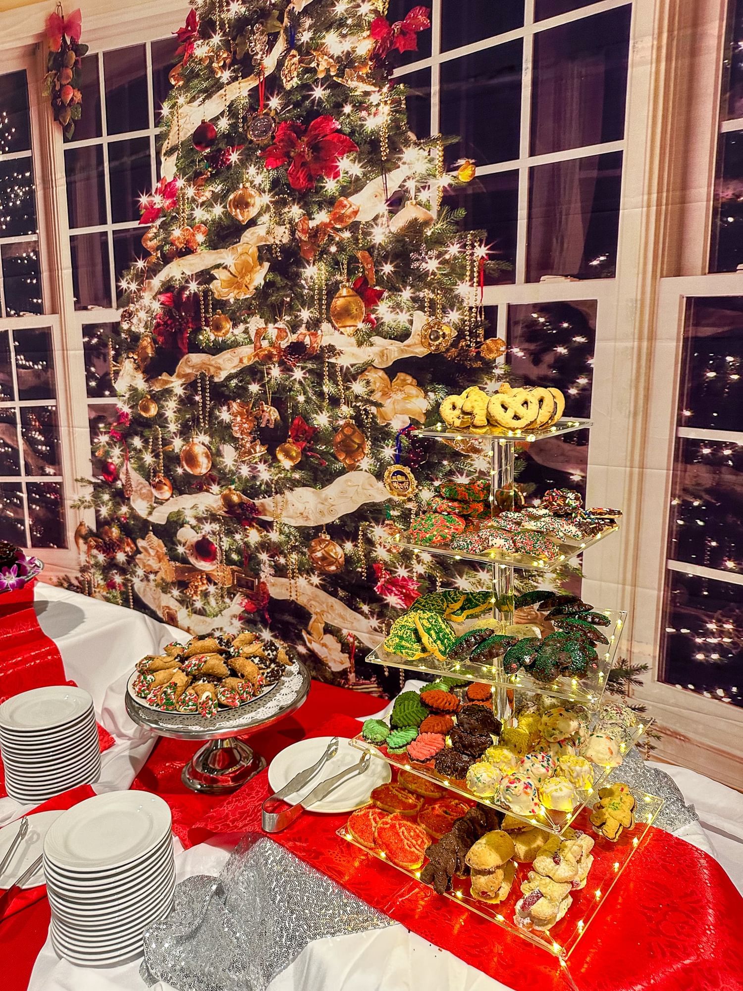 A tray of cannoli and five tiers of Christmas cookies in front of a backdrop of a sparkling Christmas tree and windows.