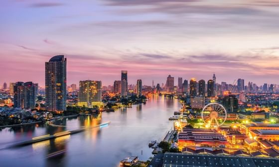 Panoramic view of Bangkok at dusk with a river, illuminated buildings, and a Ferris wheel.