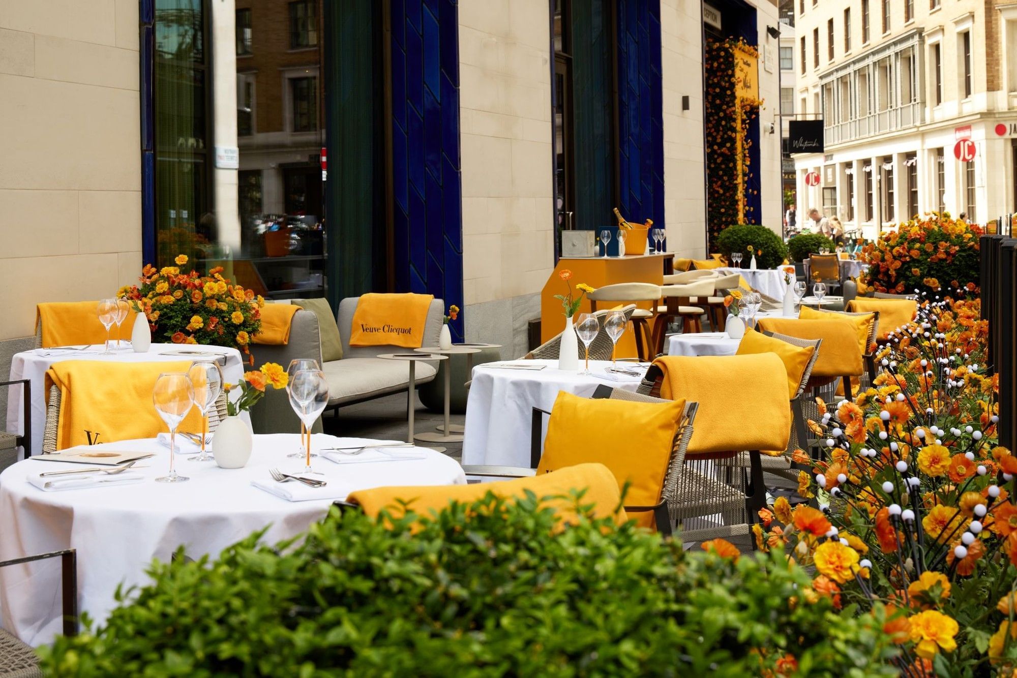 Outdoor terrace dining with white-clothed tables, yellow cushioned seating, and vibrant orange floral displays