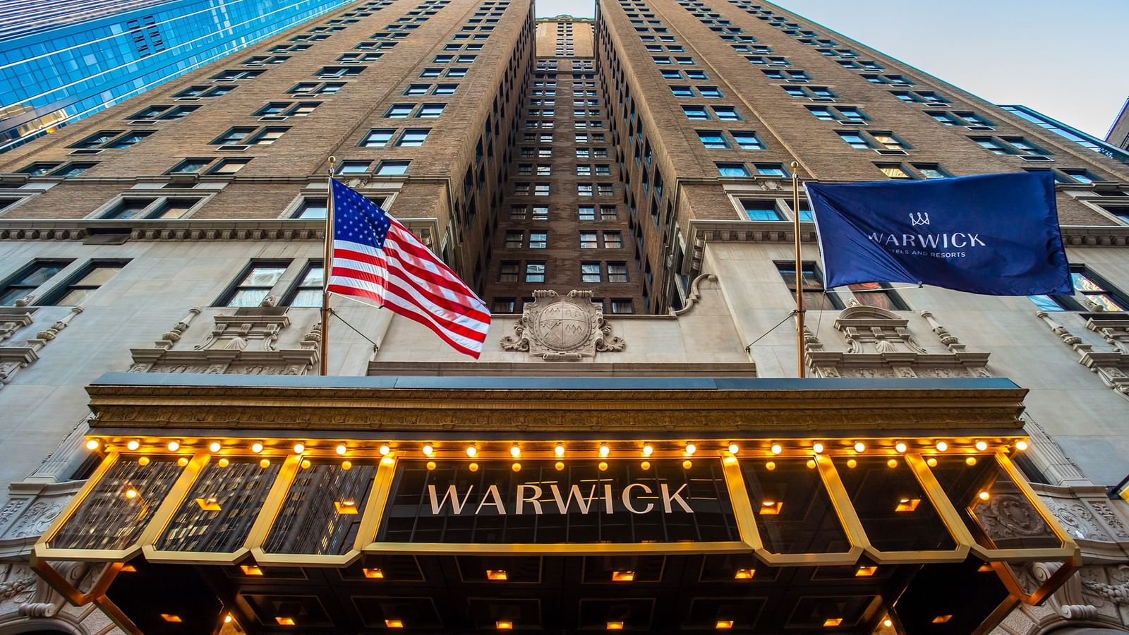 Upward view of Warwick New York's entrance with illuminated marquee, American flag, and towering facade in Midtown New York.