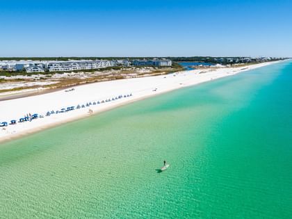 Aerial view of a pristine white sand beach with turquoise waters, blue umbrellas, and coastal buildings in the distance.