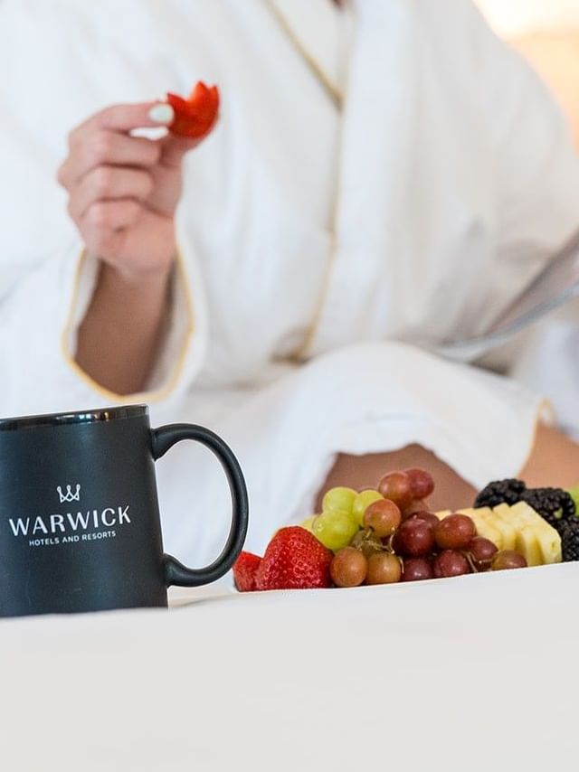 Woman in a robe enjoying a fruit platter and magazine at Warwick Member Indulgence.