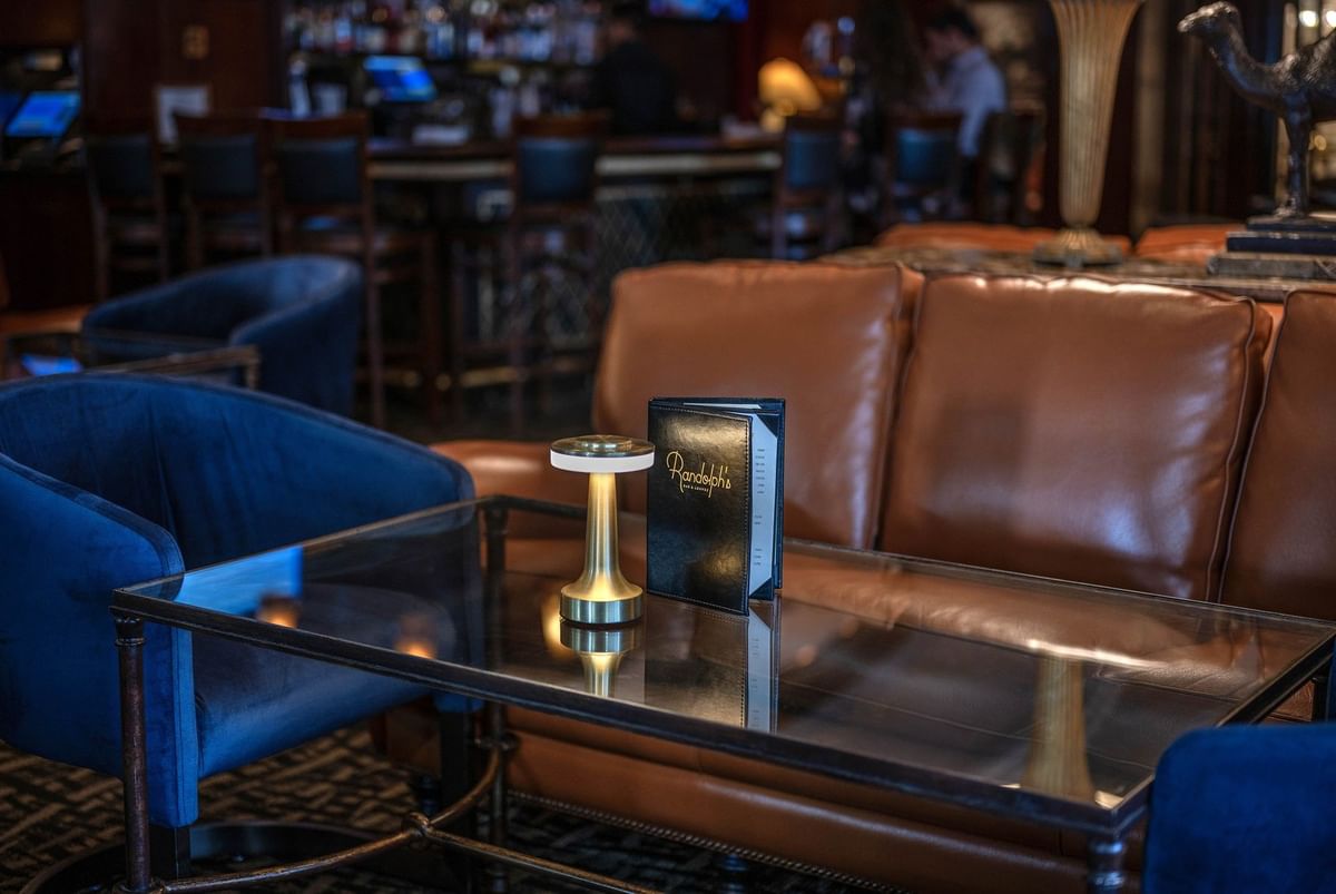 Close-up of a leather sofa and glass coffee table with a menu and gold lamp in Randolph's Bar & Lounge at Warwick New York