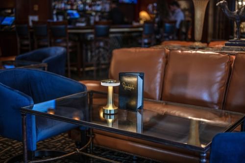 Close-up of a leather sofa and glass coffee table with a menu and gold lamp in Randolph's Bar & Lounge at Warwick New York