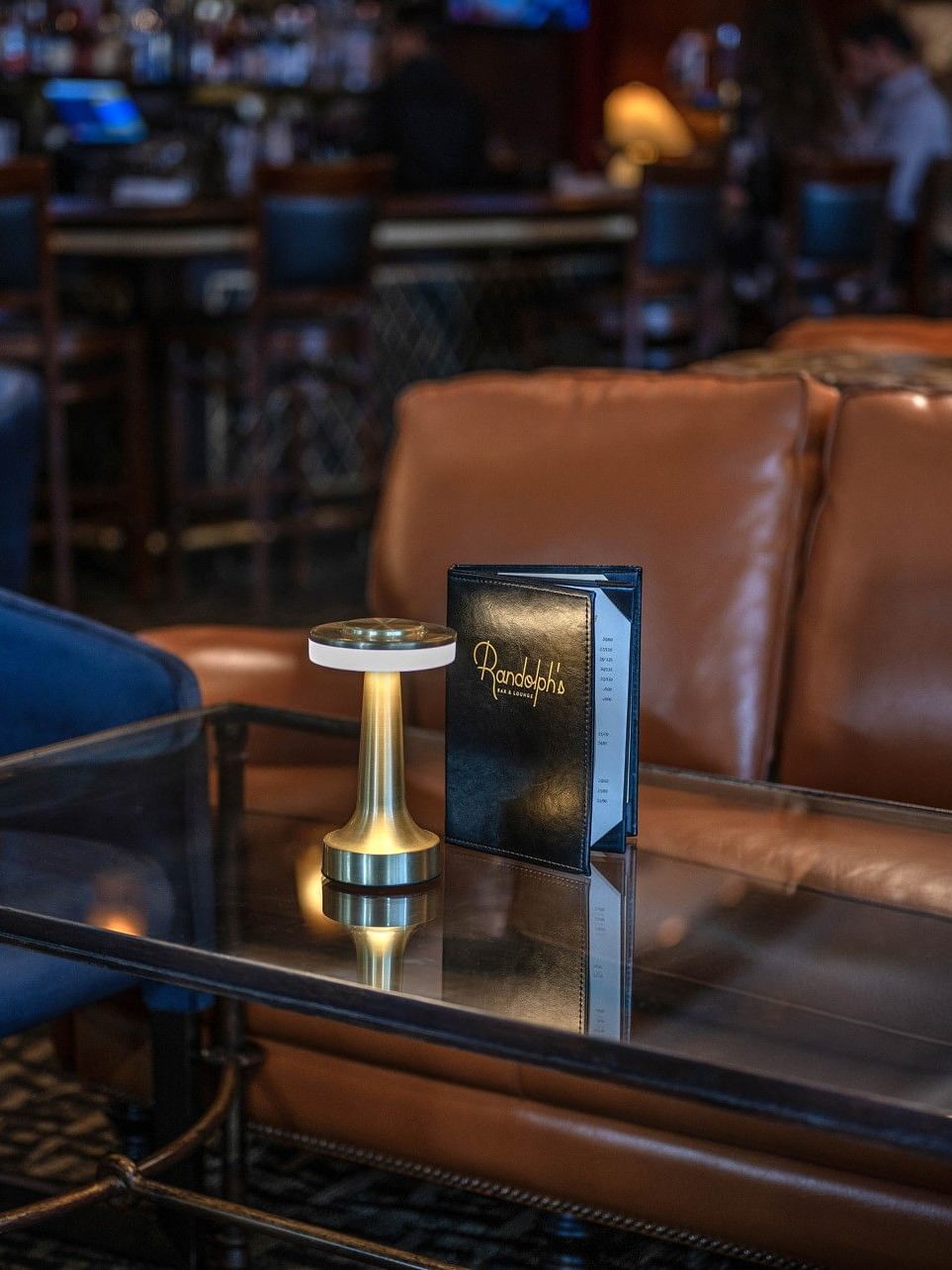 Close-up of a leather sofa and glass coffee table with a menu and gold lamp in Randolph's Bar & Lounge at Warwick New York