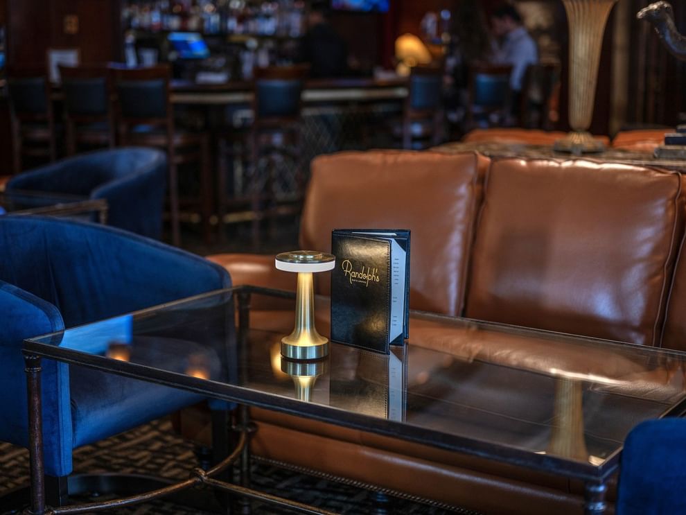 Close-up of a leather sofa and glass coffee table with a menu and gold lamp in Randolph's Bar & Lounge at Warwick New York