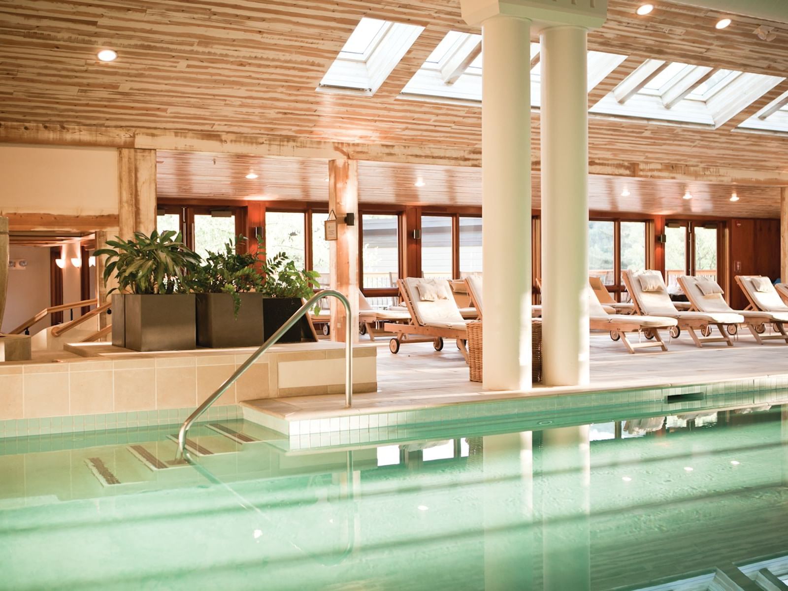Indoor pool with skylights, wooden lounge chairs, and green plants on tiled platform with handrail.