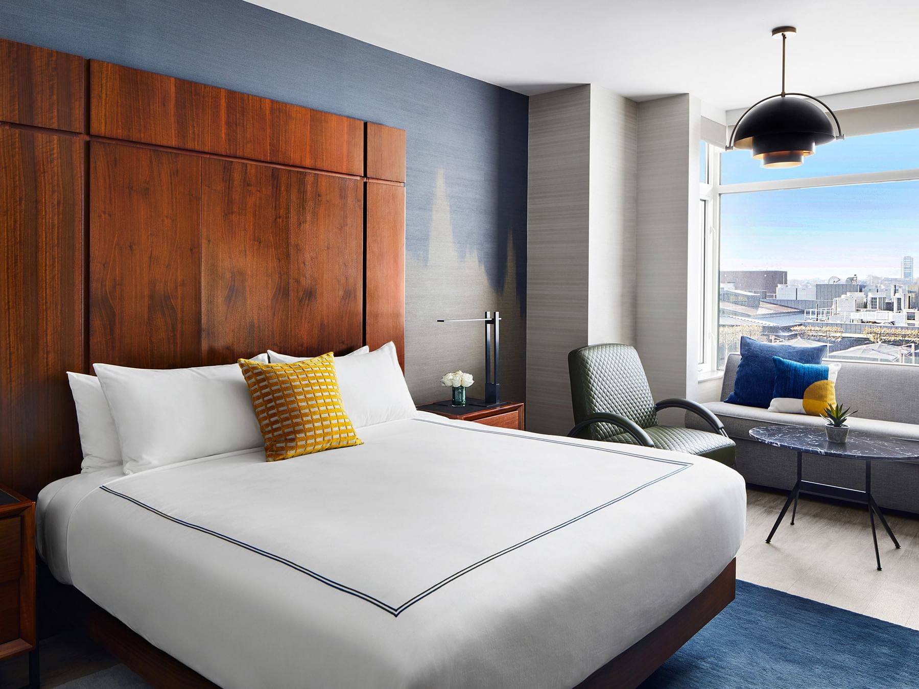 Elegant hotel room with large bed, blue couch, round table, and cityscape view from the window.
