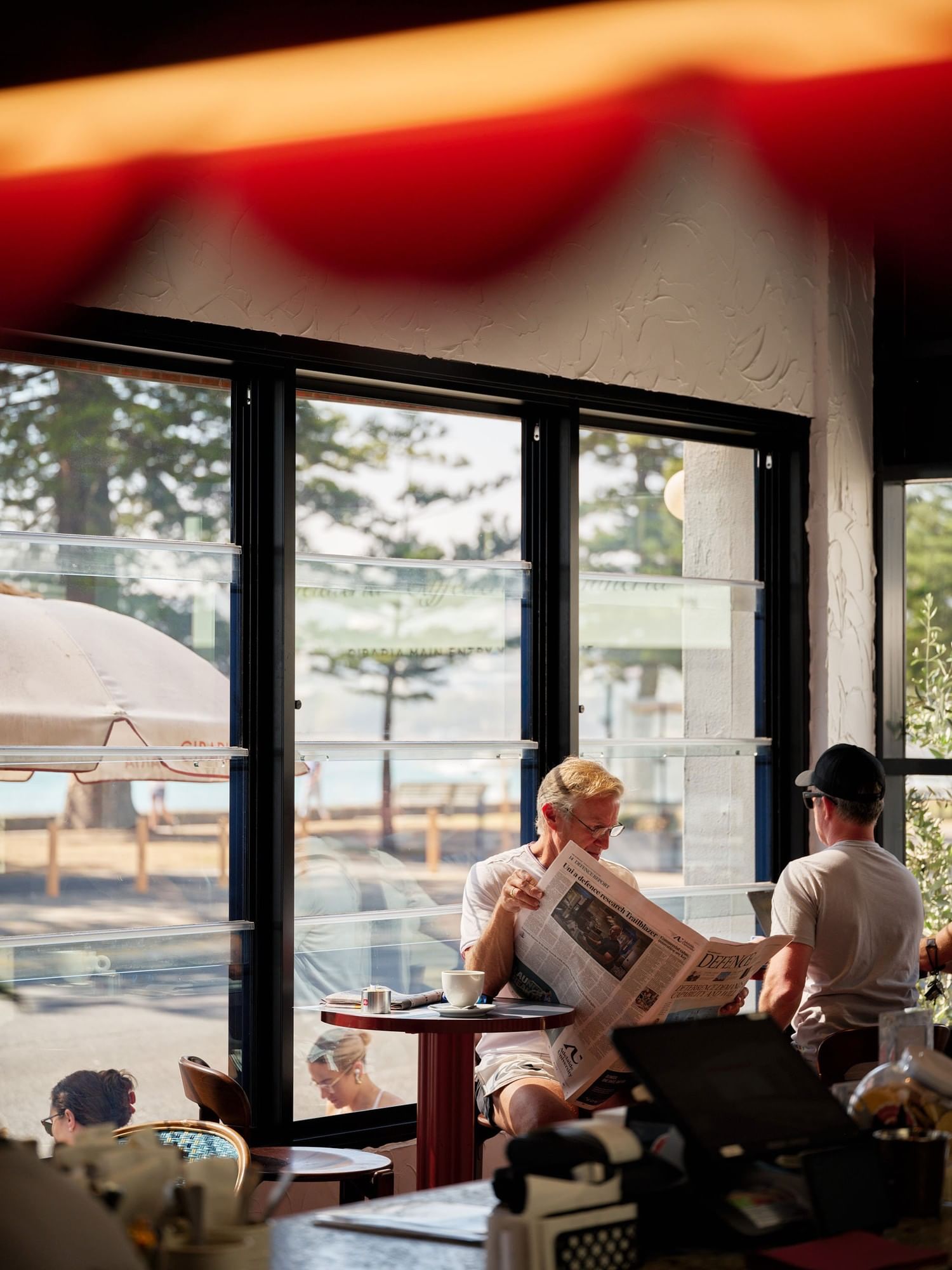 Cibaria Manly at Elysium Hotels with man reads newspaper beside another man by window with ocean and trees view