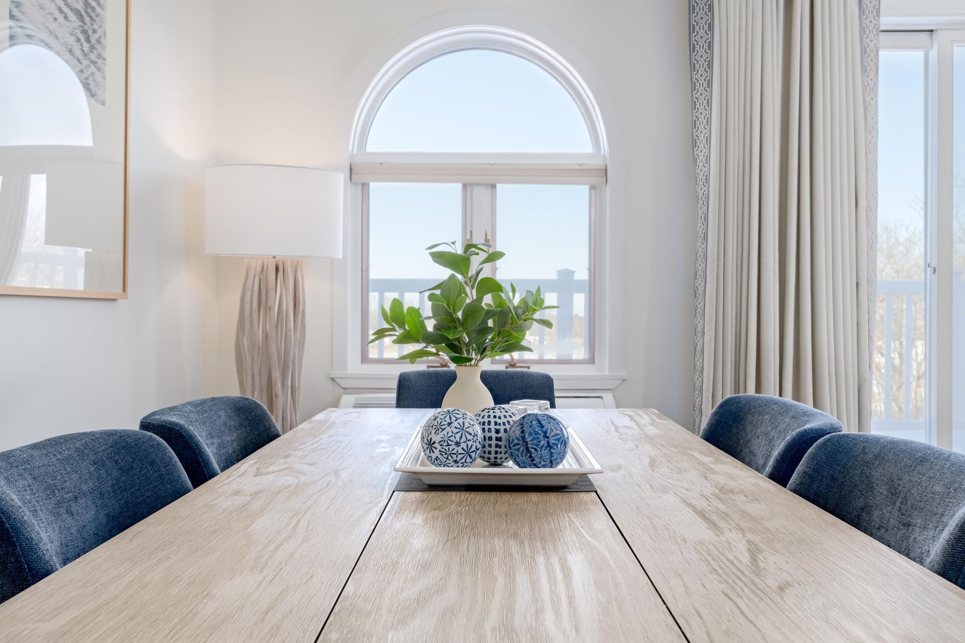 A dining room with a large wooden table, blue chairs, and a vase with greenery on the table in front of a large arched window.