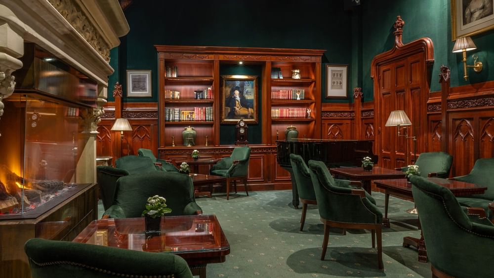 Green velvet chairs by wooden tables surrounding a grand piano and bookshelf under dark walls at Warwick Hotels & Resorts