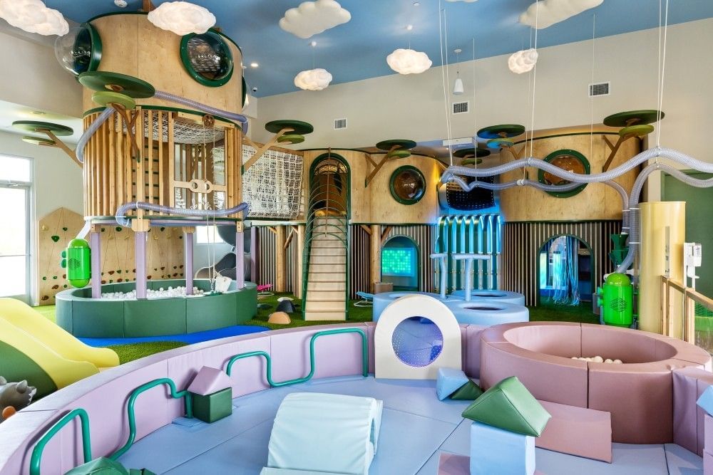 A beautifully aesthetic indoor playground with wooden towers, purple and blue climbing equipment, and a blue sky-like ceiling.