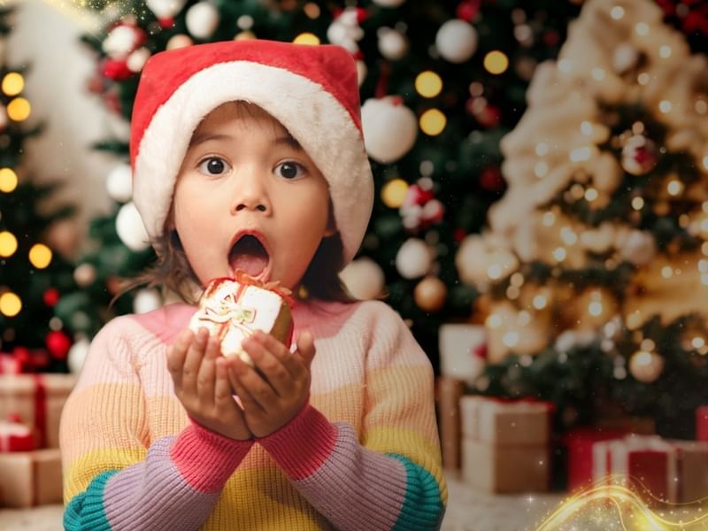 A child in a Santa hat holds a gift with mouth open, in front of decorated Christmas trees.