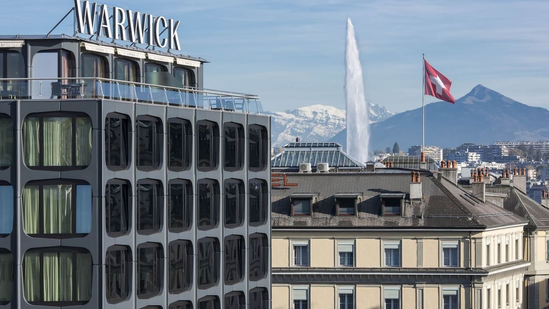 Modern Warwick Geneva Hotel exterior by the Jet d'Eau fountain under snowy mountains