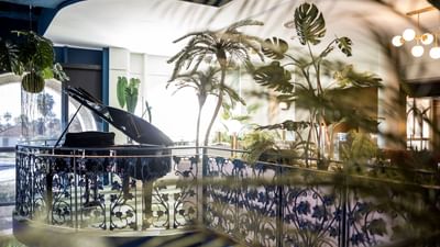 A grand piano sits on a balcony surrounded by lush greenery and a decorative railing.