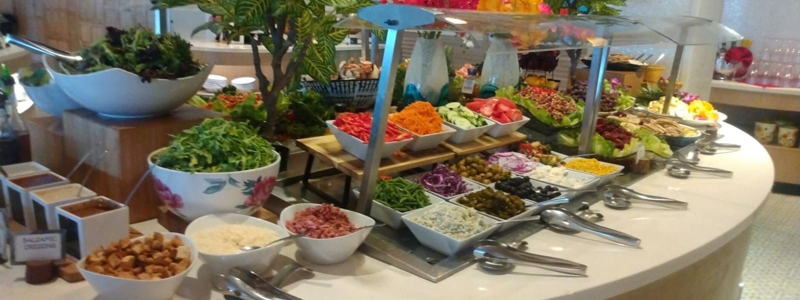 Circular buffet table with various salads, condiments, and utensils, surrounded by decorative plants and flowers