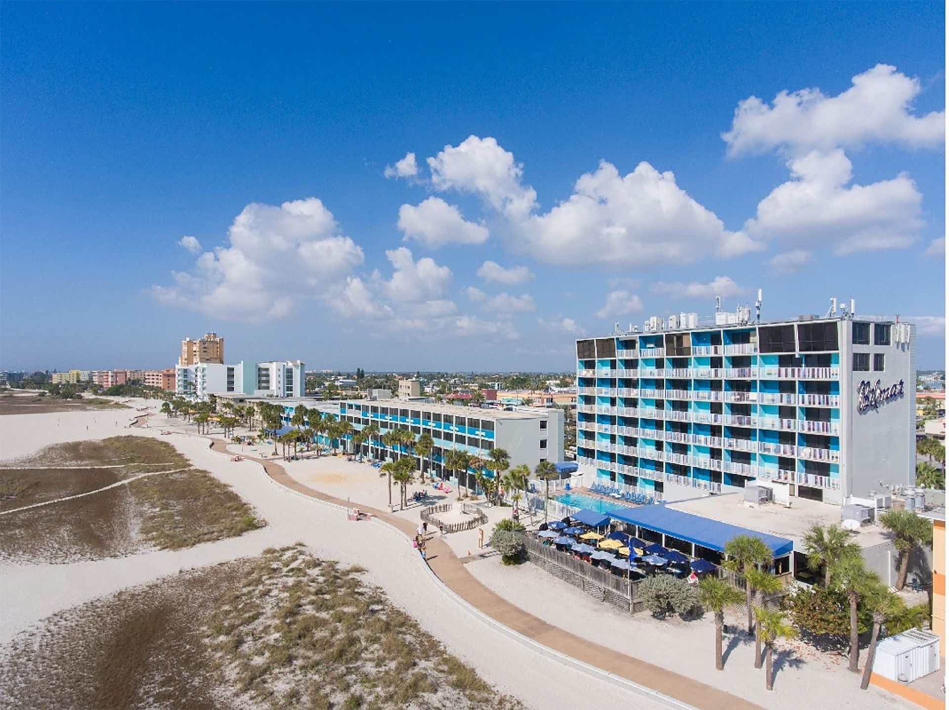 A wide-angle view of a multi-story hotel with blue and white balconies, situated on a beach with a pool and palm trees.