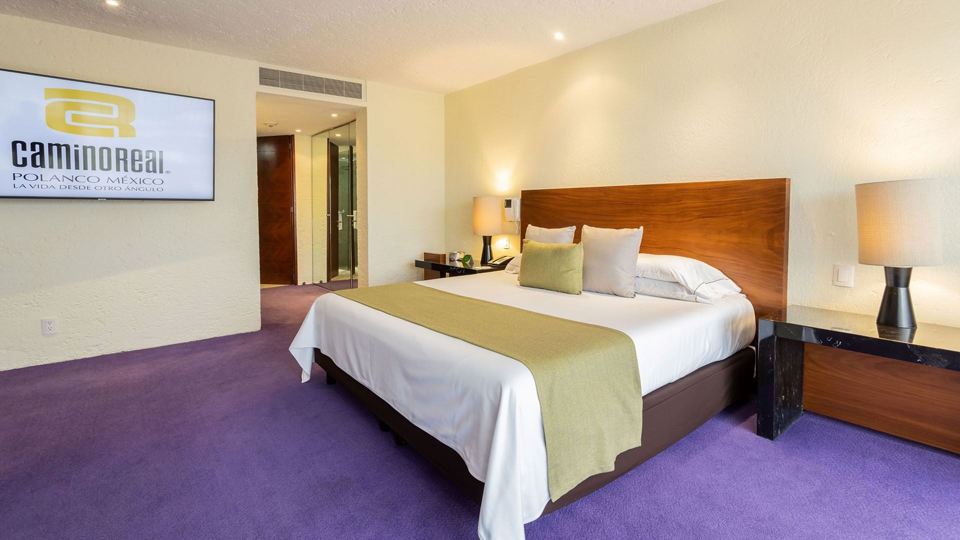 Habitación de hotel con cama grande, pantalla en la pared y alfombra púrpura.