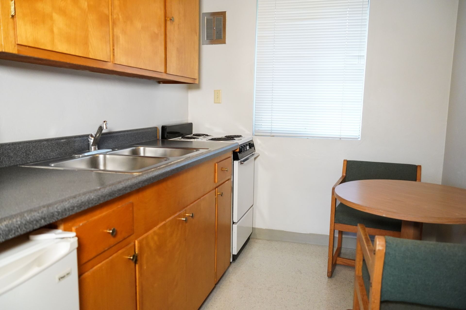 A compact kitchenette with wood cabinets, gray countertop, and a small dining area with a round table and two chairs.