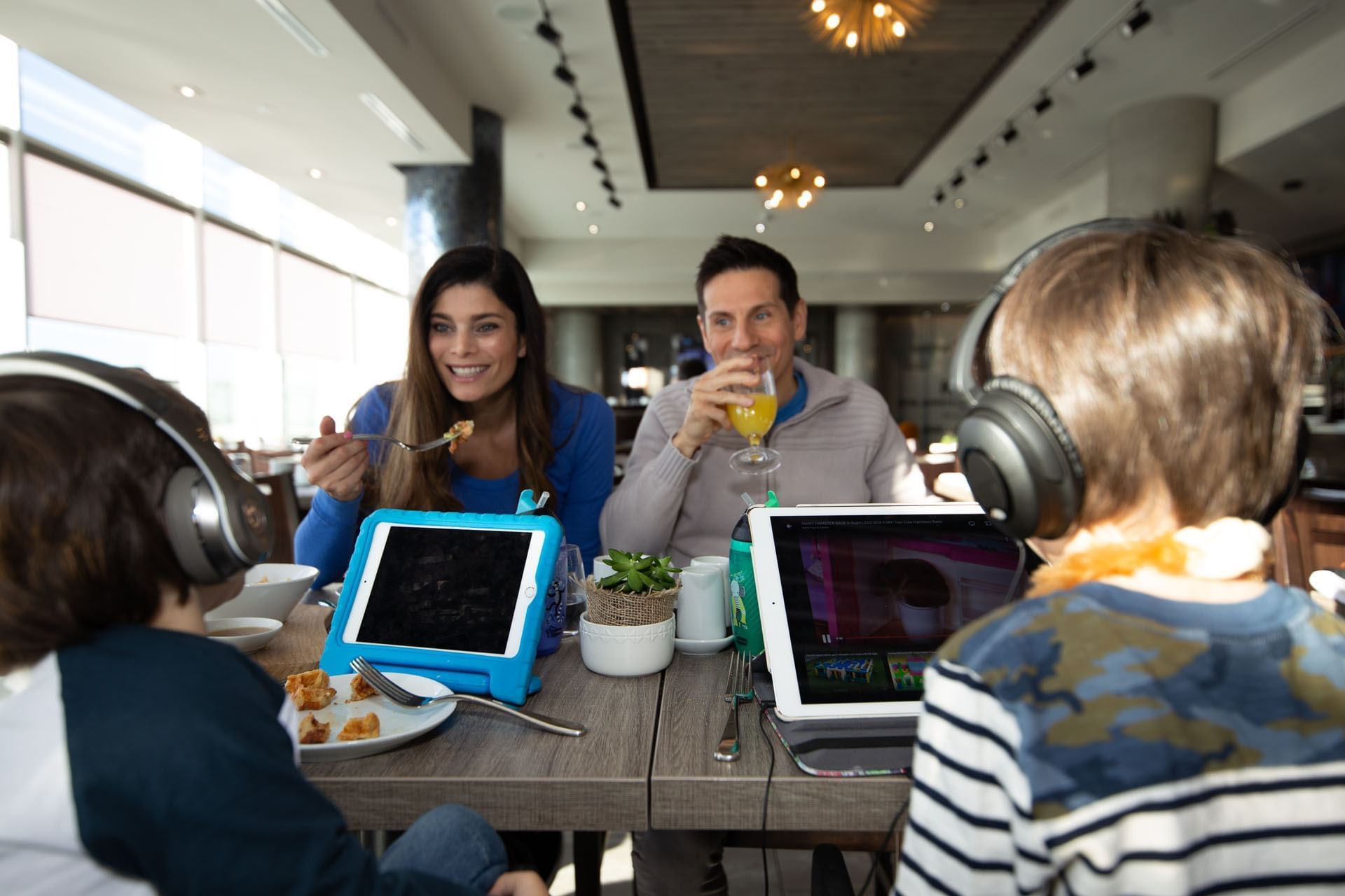 Family with tablets and headphones by a wooden table, eating brunch under warm pendant lighting at the Hotel X Toronto