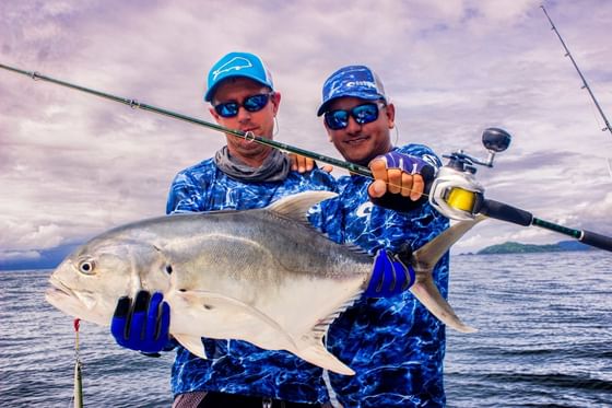 Two smiling men holding a large jack fish with a fishing rod and reel on the open ocean near Jungle Vista Boutique Hotel