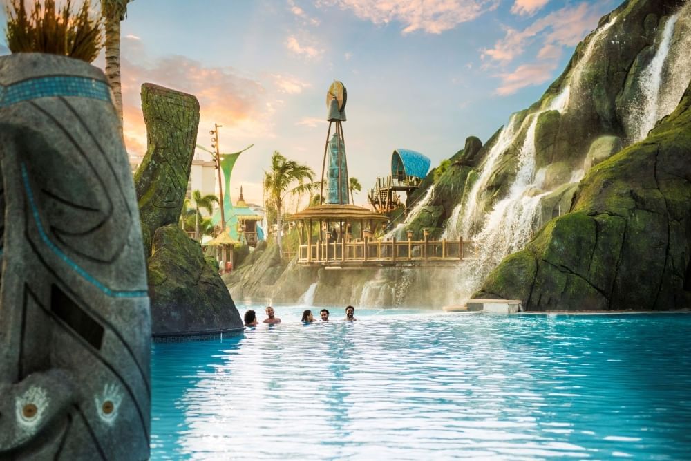 Five people swim in the distance of a grotto surrounded by stone walls and the side of a volcano with a waterfall. 