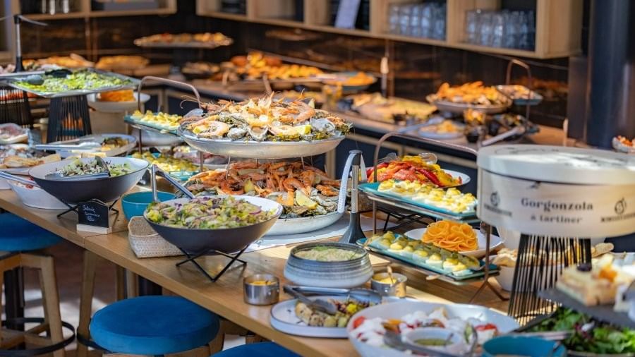 A buffet table laden with various seafood and salad dishes, with blue stools positioned around it.