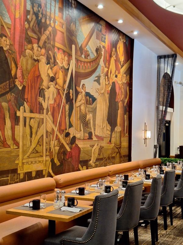 Long dining table with orange banquette seating positioned before large historical mural in Murals on 54 at Warwick New York