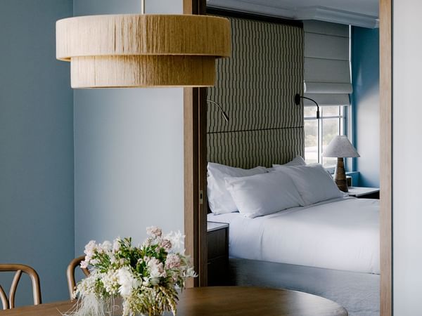 Floral centerpiece on a round table at Elysium Noosa beside a woven chair, with a plush white bed framed in the doorway